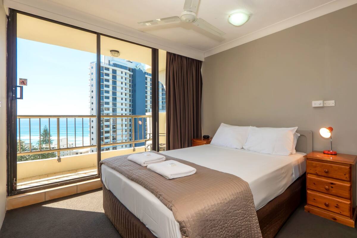 A hotel room with a large bed, a bedside table with a lamp, a wooden dresser, and a view of tall buildings and the ocean.