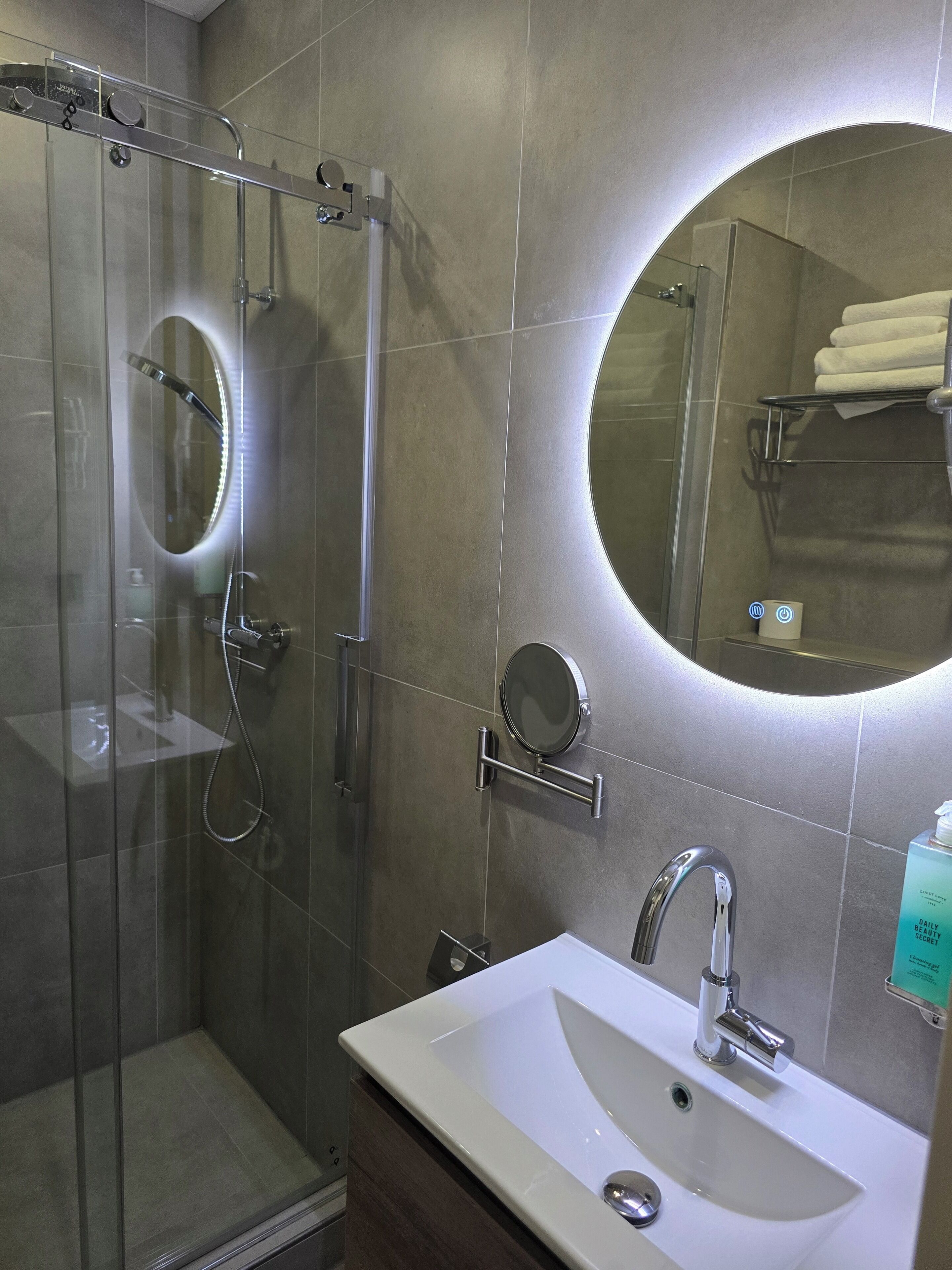 A modern bathroom with a glass-enclosed shower, a round mirror, a white sink, and a towel rack.