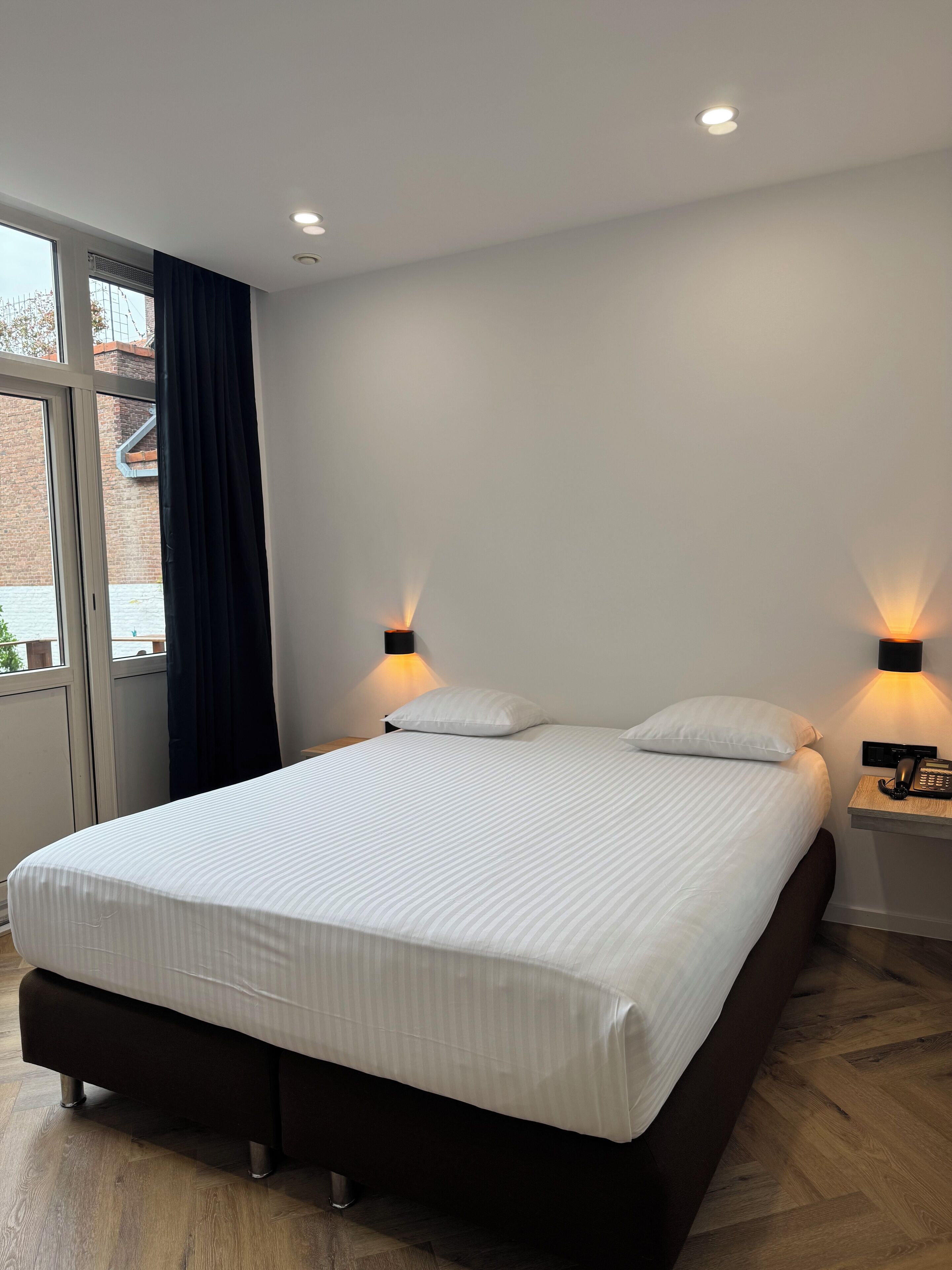 A modern hotel room with a large bed, two bedside lamps, a nightstand with a phone, and a window with curtains.