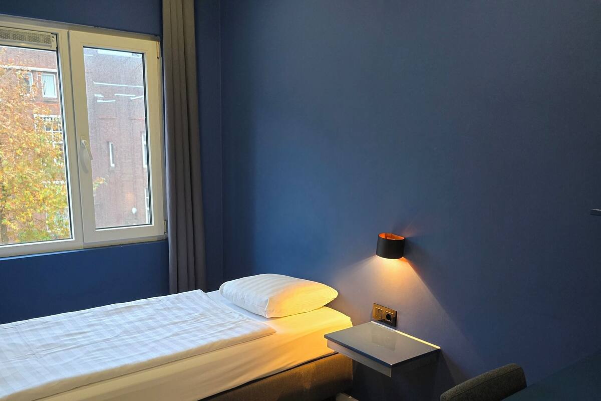 Um quarto com cama, escrivaninha, cadeira, mesa de cabeceira e janela.