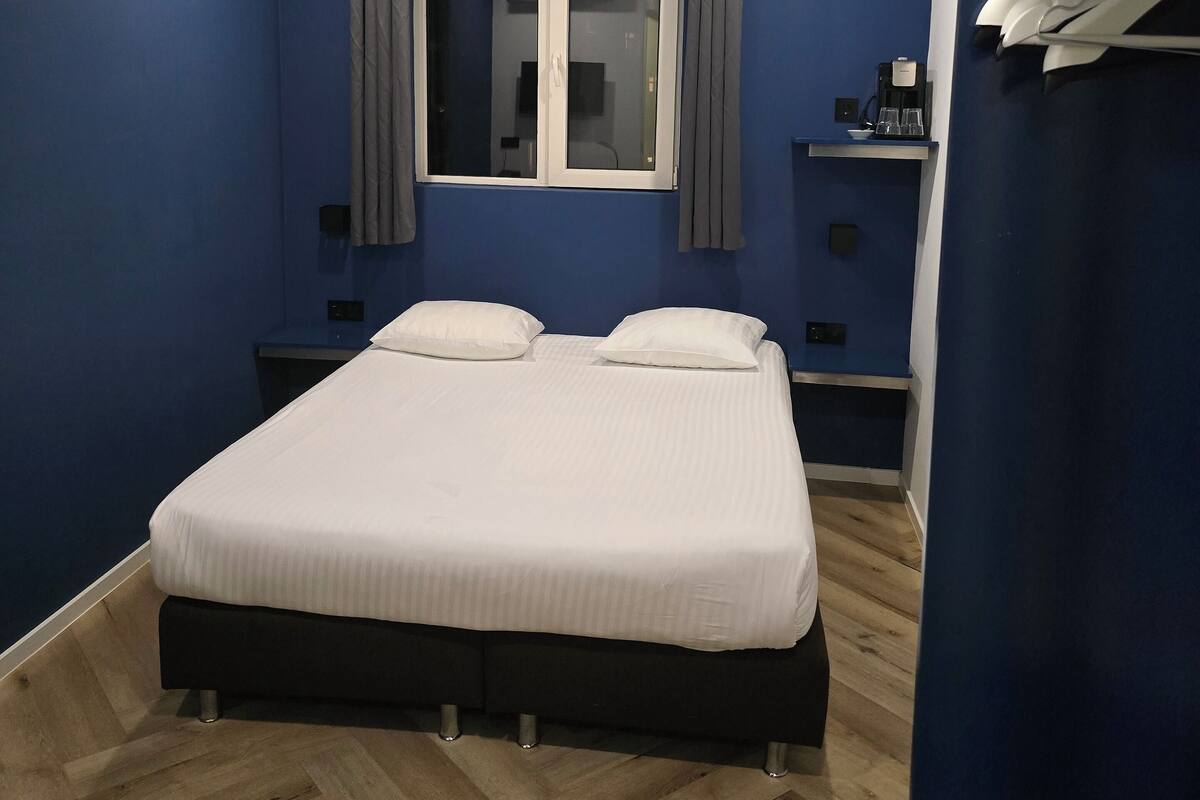 Quarto com cama, duas mesas de cabeceira, janela com cortinas e prateleira com cafeteira e copos.