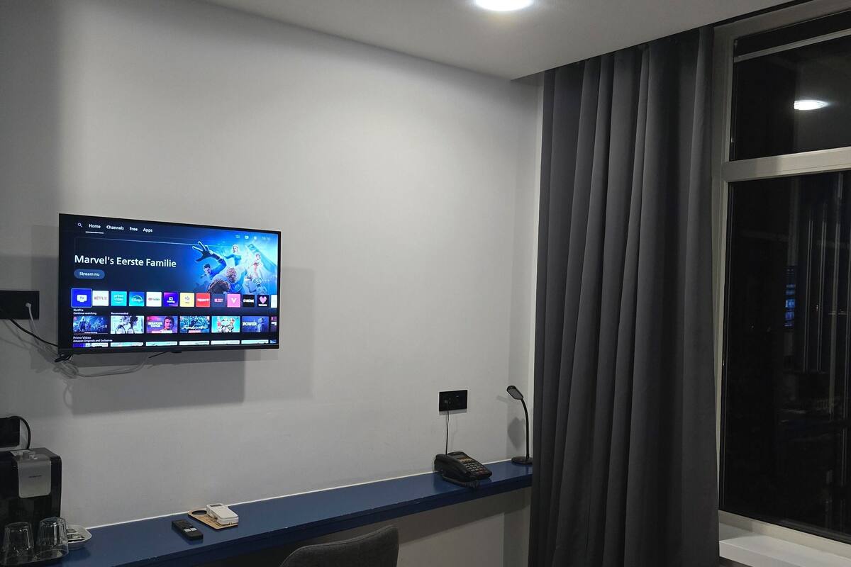 Quarto de hotel com uma cama, uma mesa com computador, uma televisão na parede, uma janela com cortinas e uma lixeira.