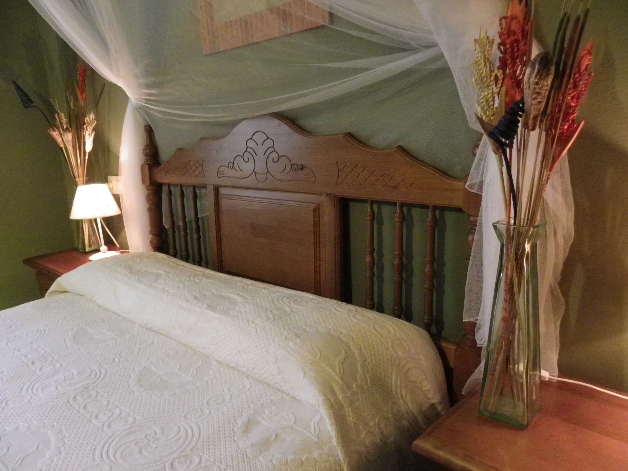 Una cama bien hecha con colcha blanca, cabecera de madera con diseño tallado, lámpara de noche y un florero con flores secas.