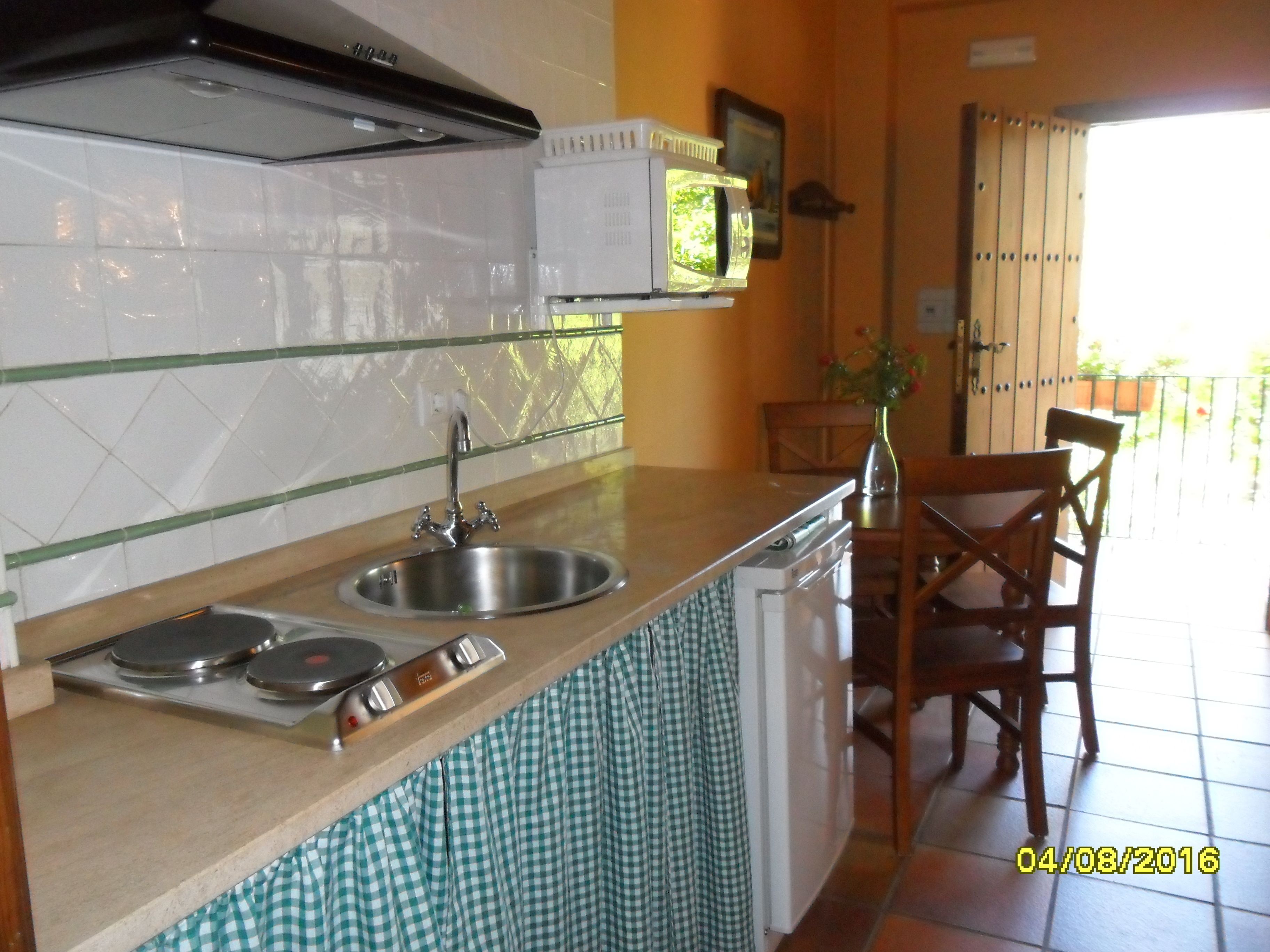 Una cocina con un lavaplatos de acero inoxidable, un microondas y una estufa. Hay una silla de madera y una mesita con un florero.