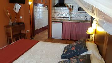 Un dormitorio con cama, almohadas, una mesita de noche, lámpara, televisor montado en la pared y vista a la cocina.