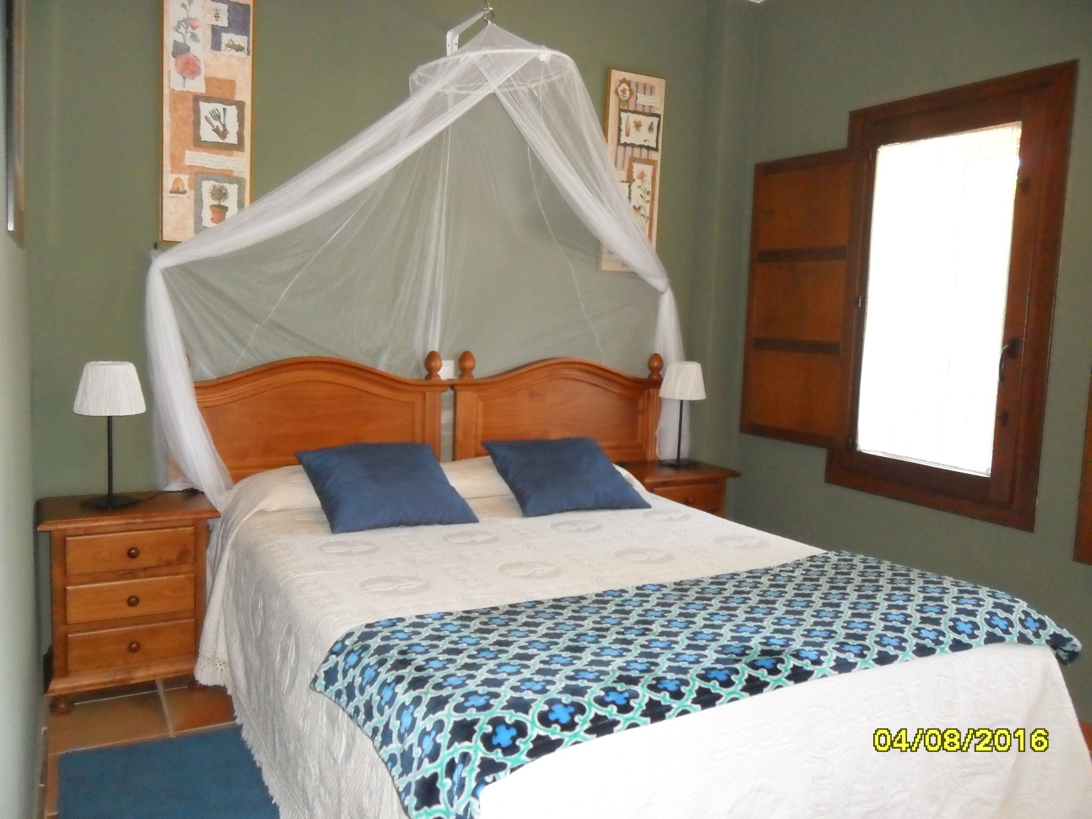 Un dormitorio con una cama de madera, dos mesitas de noche, una lámpara de noche, un ventanal y una pared decorada con marcos.