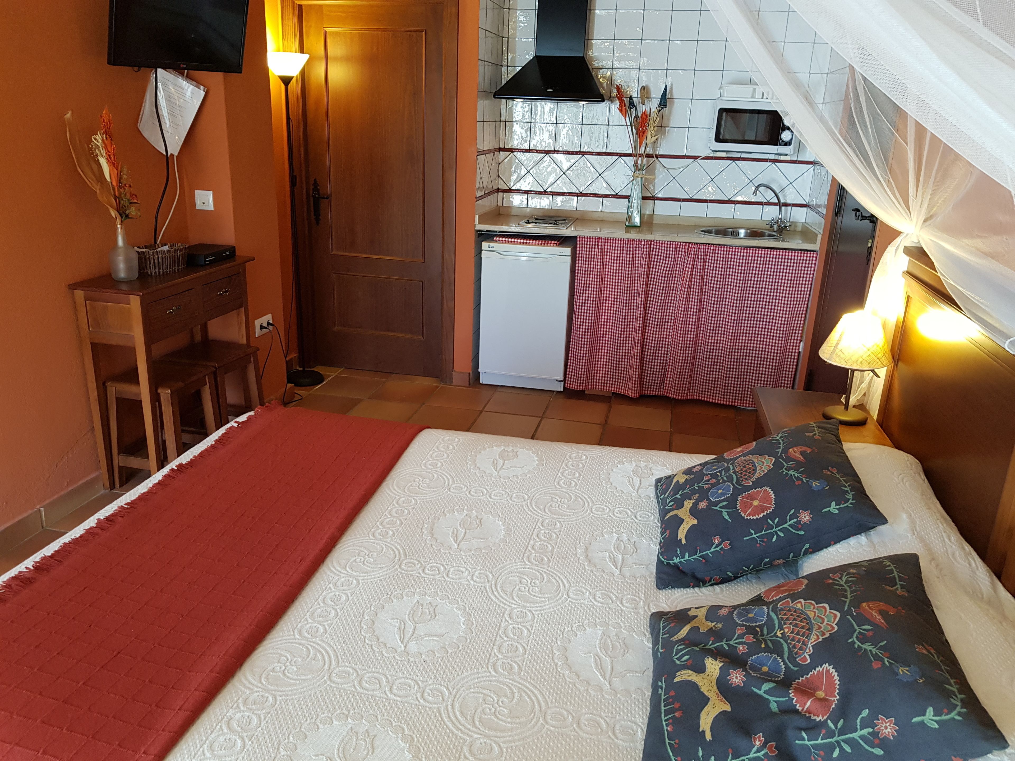 Ein kleines Hotelzimmer mit Bett, Schreibtisch, einer Küchenzeile und einem an der Wand befestigten Fernseher.