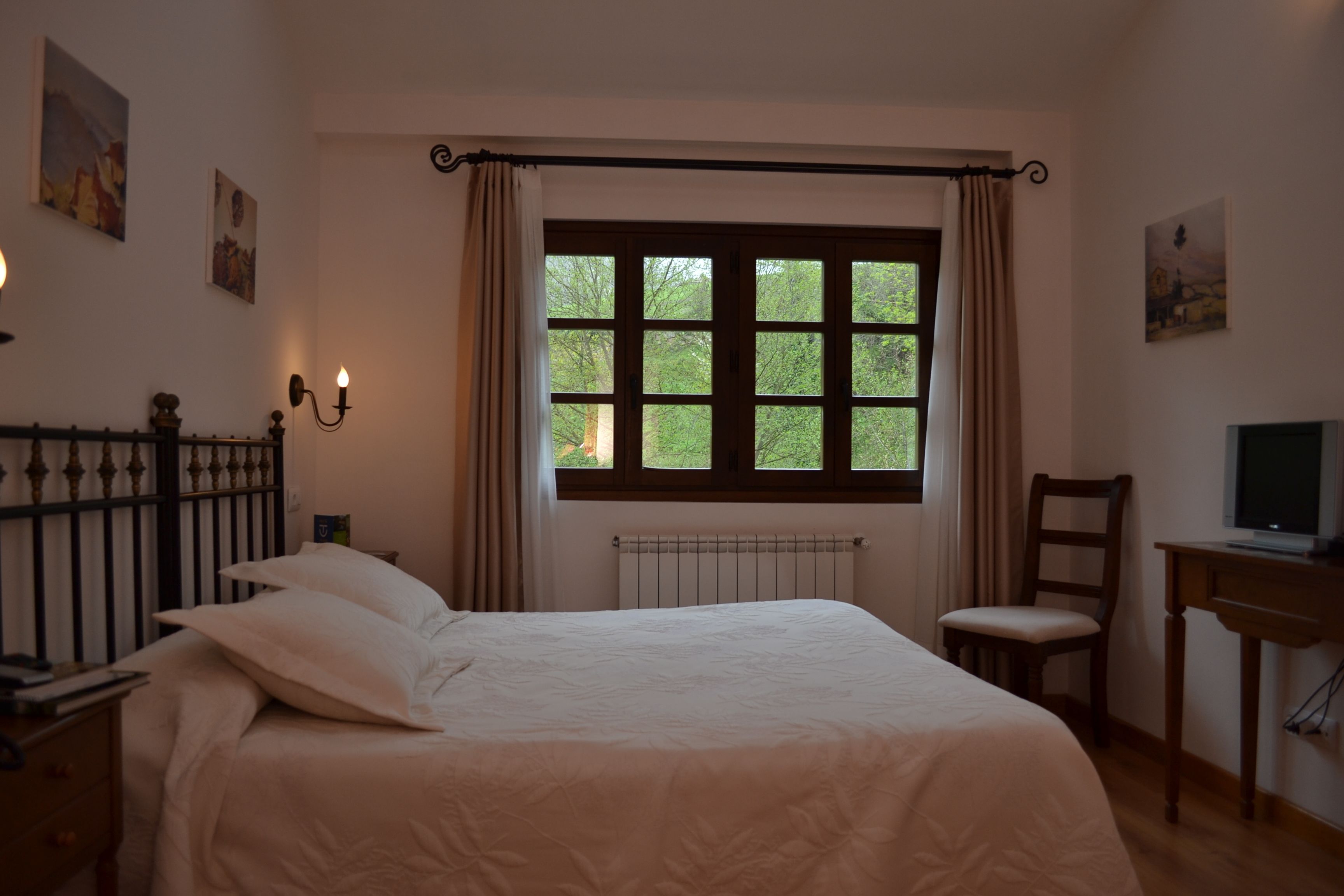 A bedroom with a large bed, a chair, a desk, a radiator, and a window with curtains.