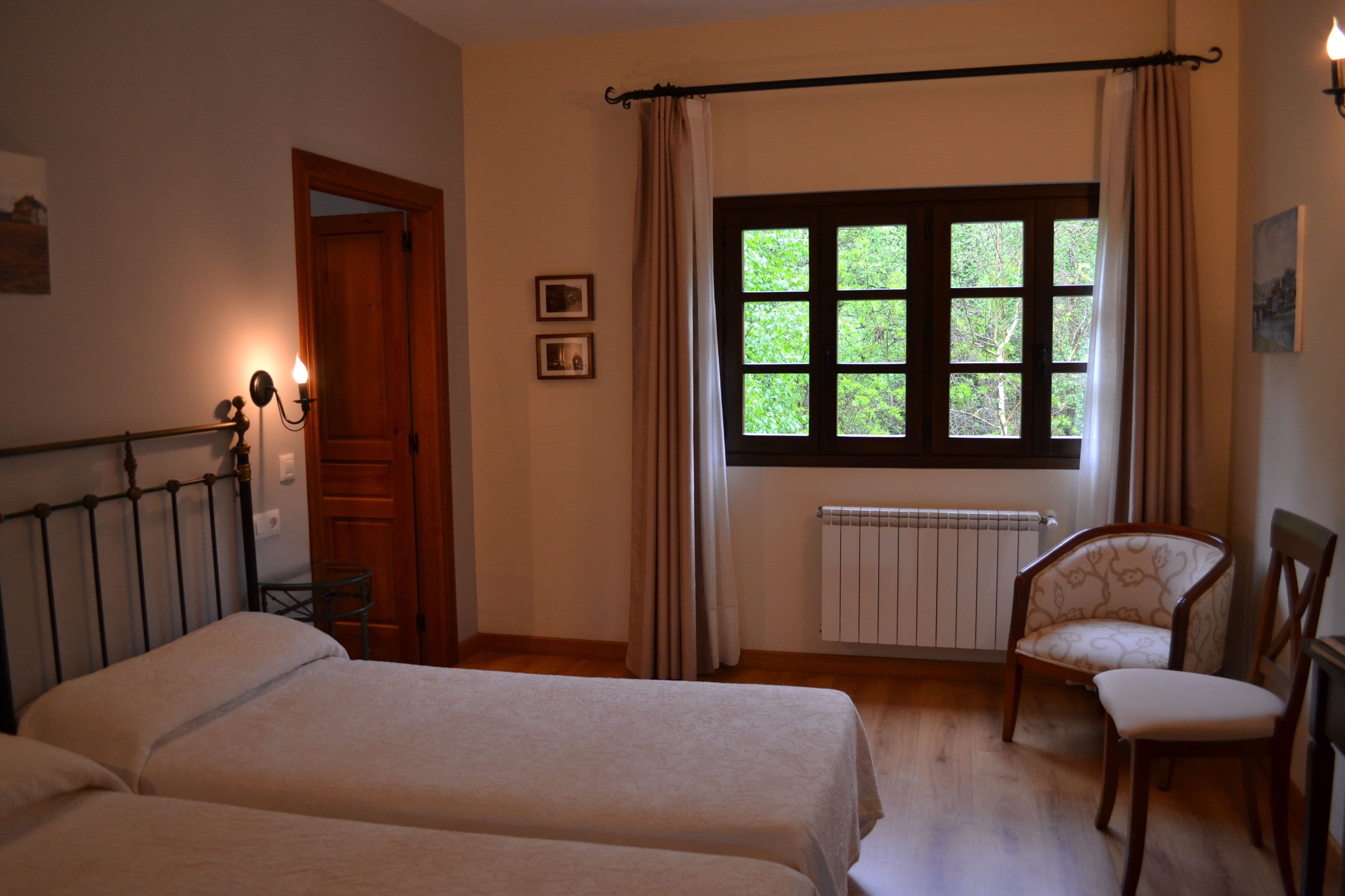 Ein Schlafzimmer mit zwei Betten, einem Sessel, einem Fenster mit Vorhängen, einem Heizkörper und einer Tür.