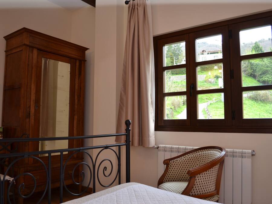 A bedroom with a bed, a wooden wardrobe, a window with a view of greenery, and a radiator.