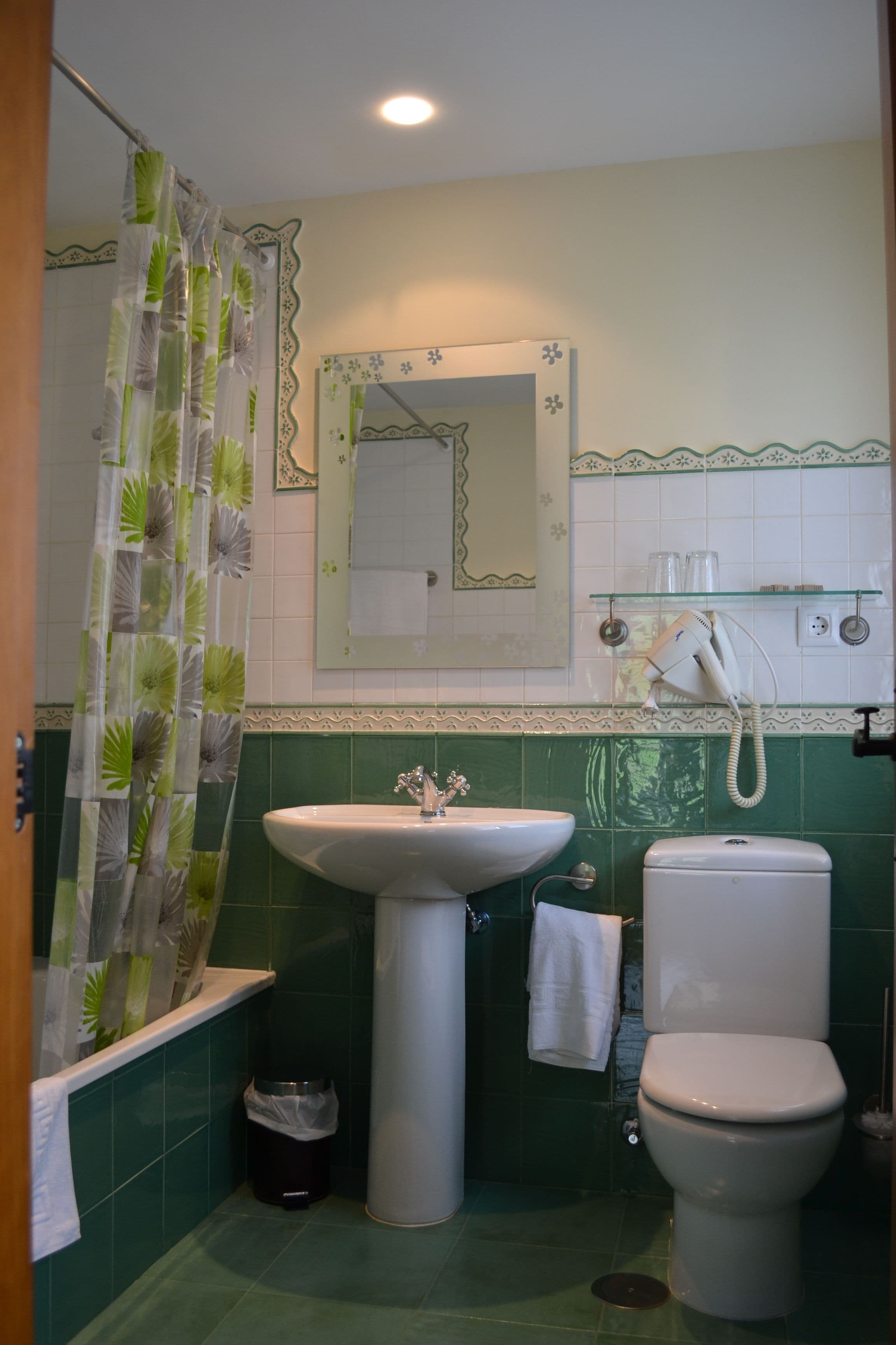 Un baño con pared de azulejos verdes, un lavabo blanco, un inodoro, una cortina de ducha y un espejo.