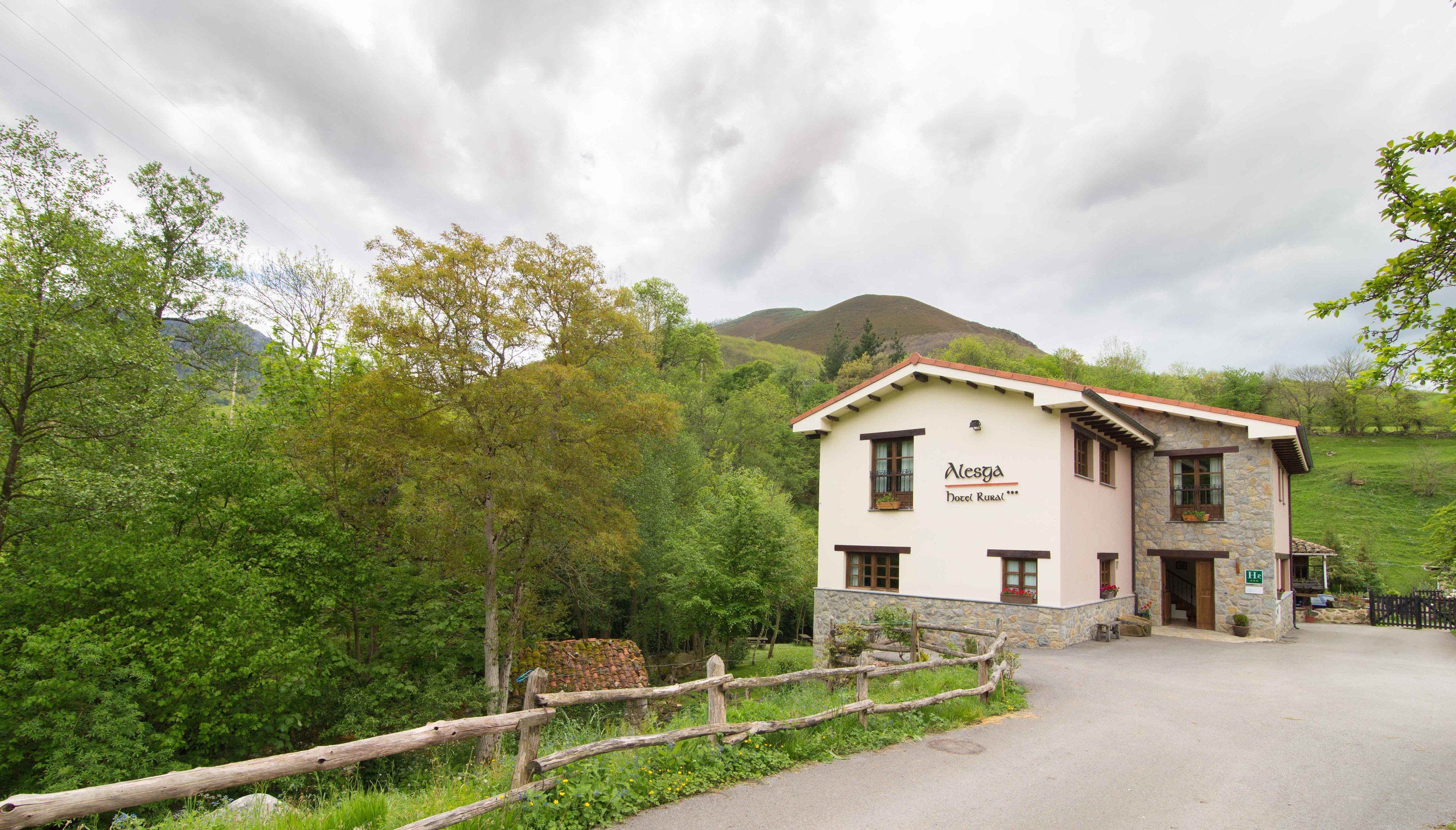 A two-story building with a sign reading "Mesga Hotel Rural." It features a stone facade, a wooden fence in front, and is surrounded by lush greenery and hills in the background.