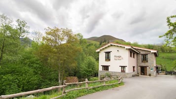 Un bâtiment à deux étages, sur lequel on peut lire l’inscription « Mesga Hotel Rural ». Il se distingue par sa façade en pierre, une clôture en bois à l’avant, et est entouré d’une végétation luxuriante, avec des collines en arrière-plan.