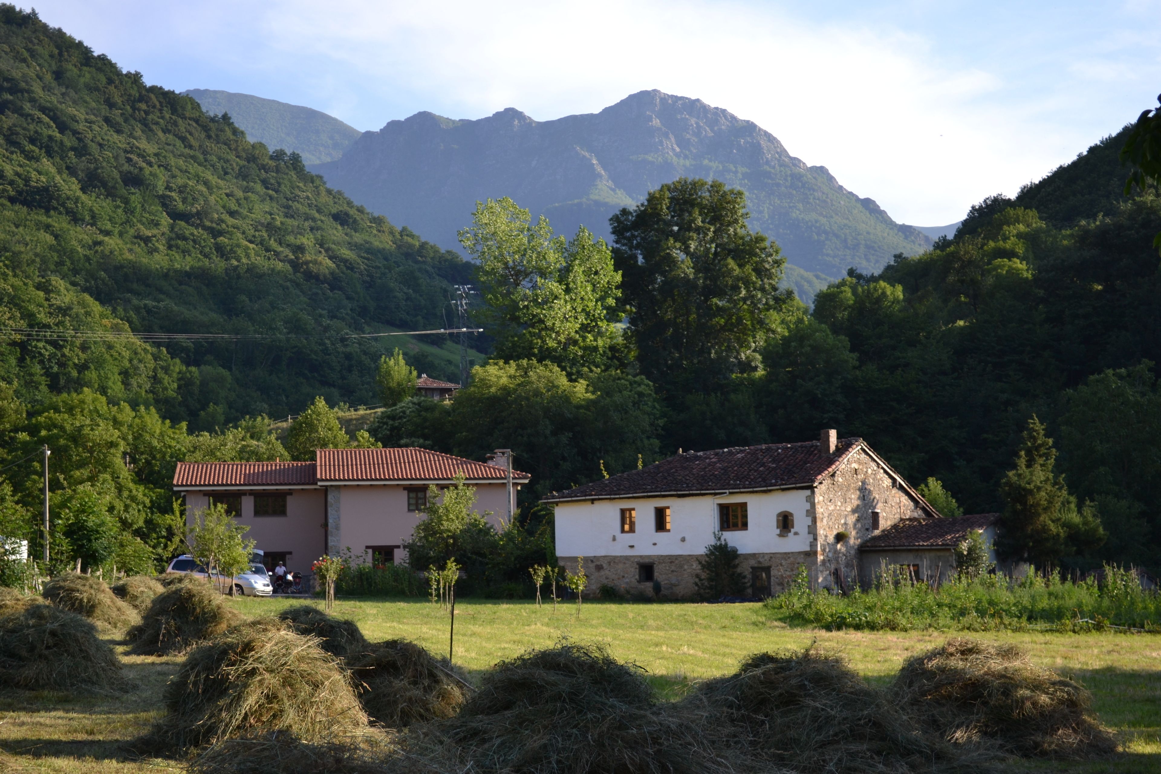 Entorno rural con dos edificios, uno con techo rojo y otro de fachada de piedra, rodeados de pacas de heno y follaje, con montañas al fondo.