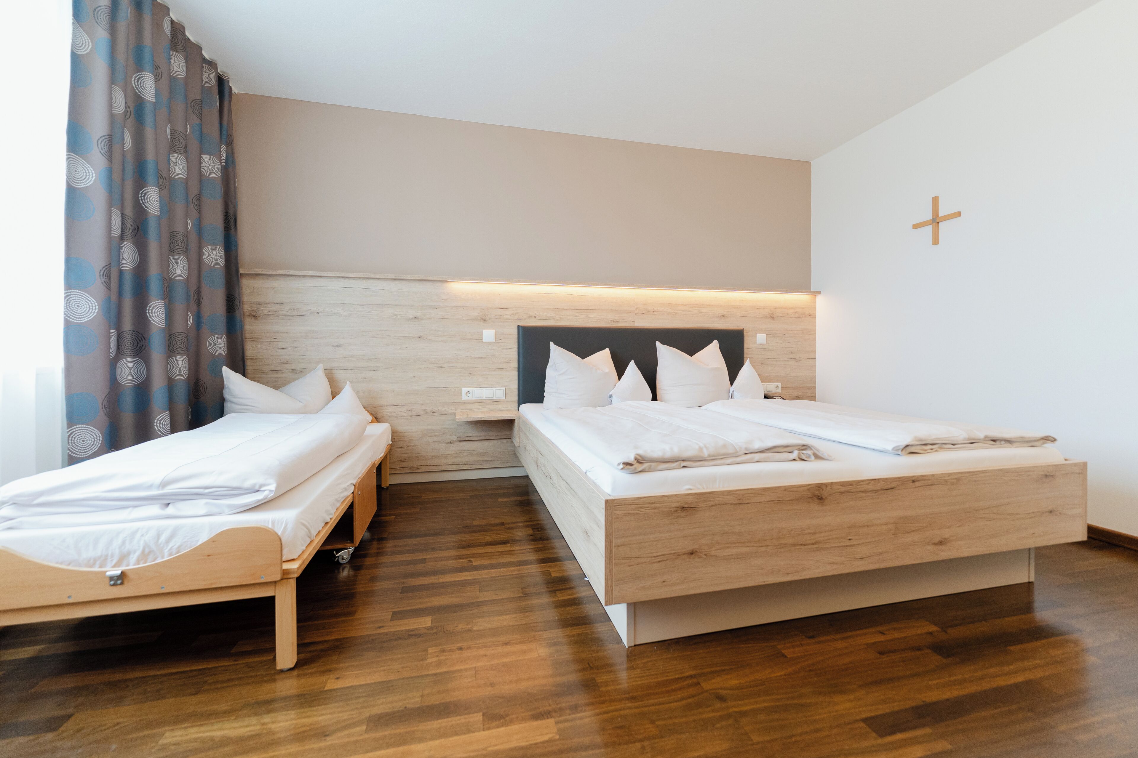 A hotel room with two single beds, a wooden headboard, a cross on the wall, and a curtain with a circular pattern.