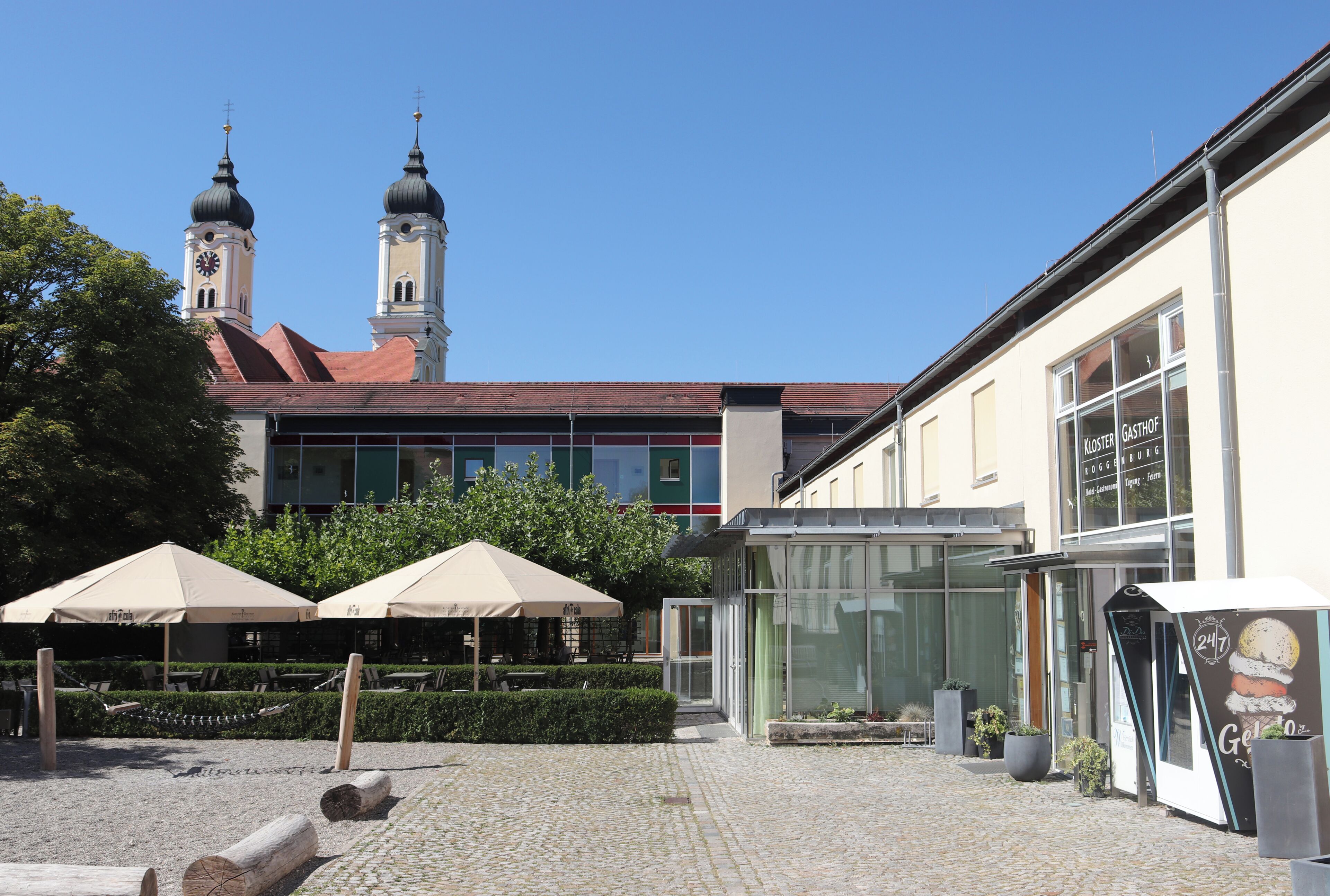 A courtyard with a church in the background, a building with a sign for 'Kloster Gasthof Roggenburg', and outdoor seating with umbrellas.