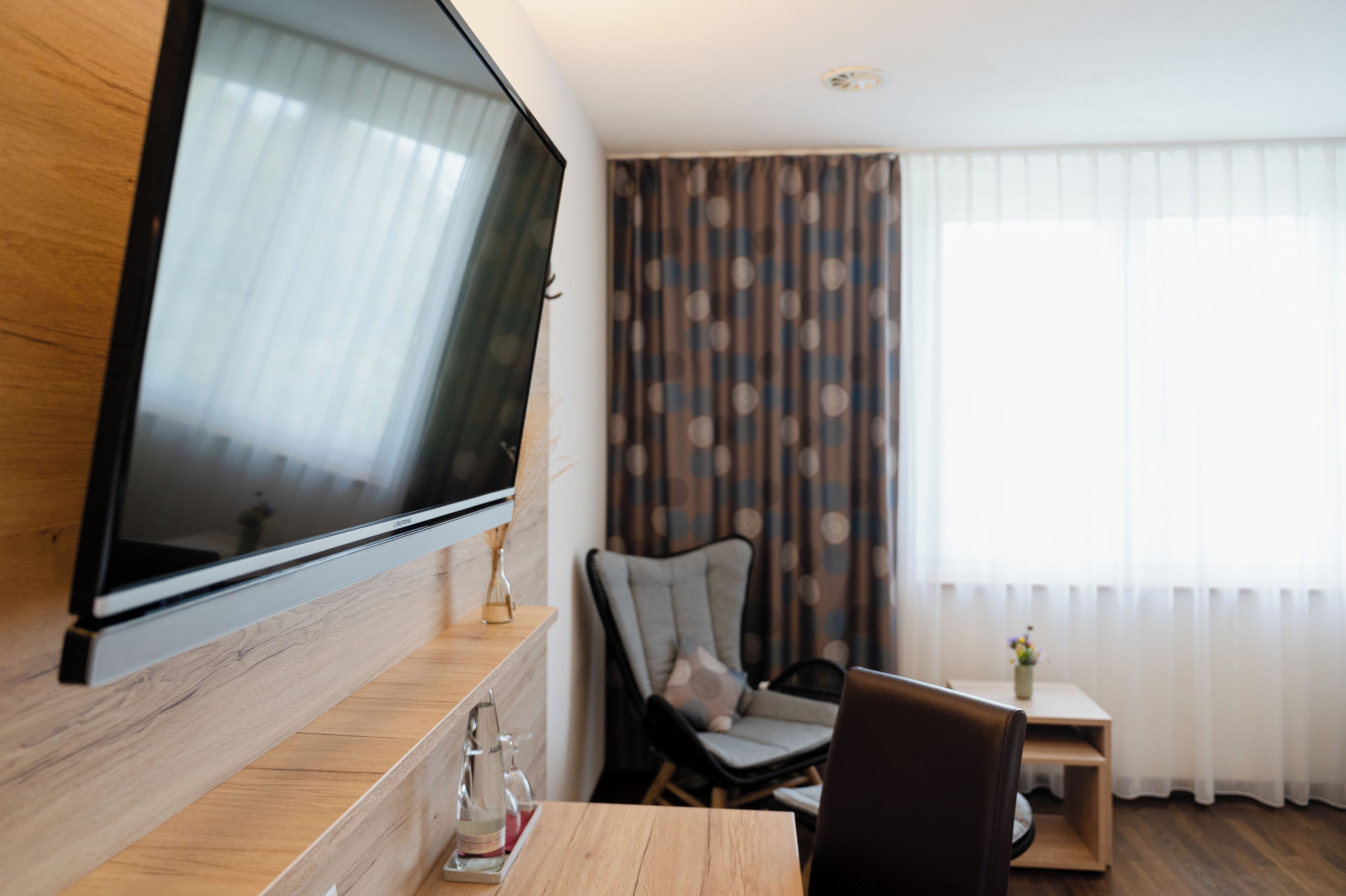 Una habitación moderna con una televisión grande montada en la pared, una mesa de madera, una silla, una mesita auxiliar y una ventana con cortinas.