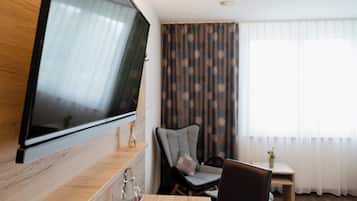 Una habitación moderna con una televisión grande montada en la pared, una mesa de madera, una silla, una mesita auxiliar y una ventana con cortinas.