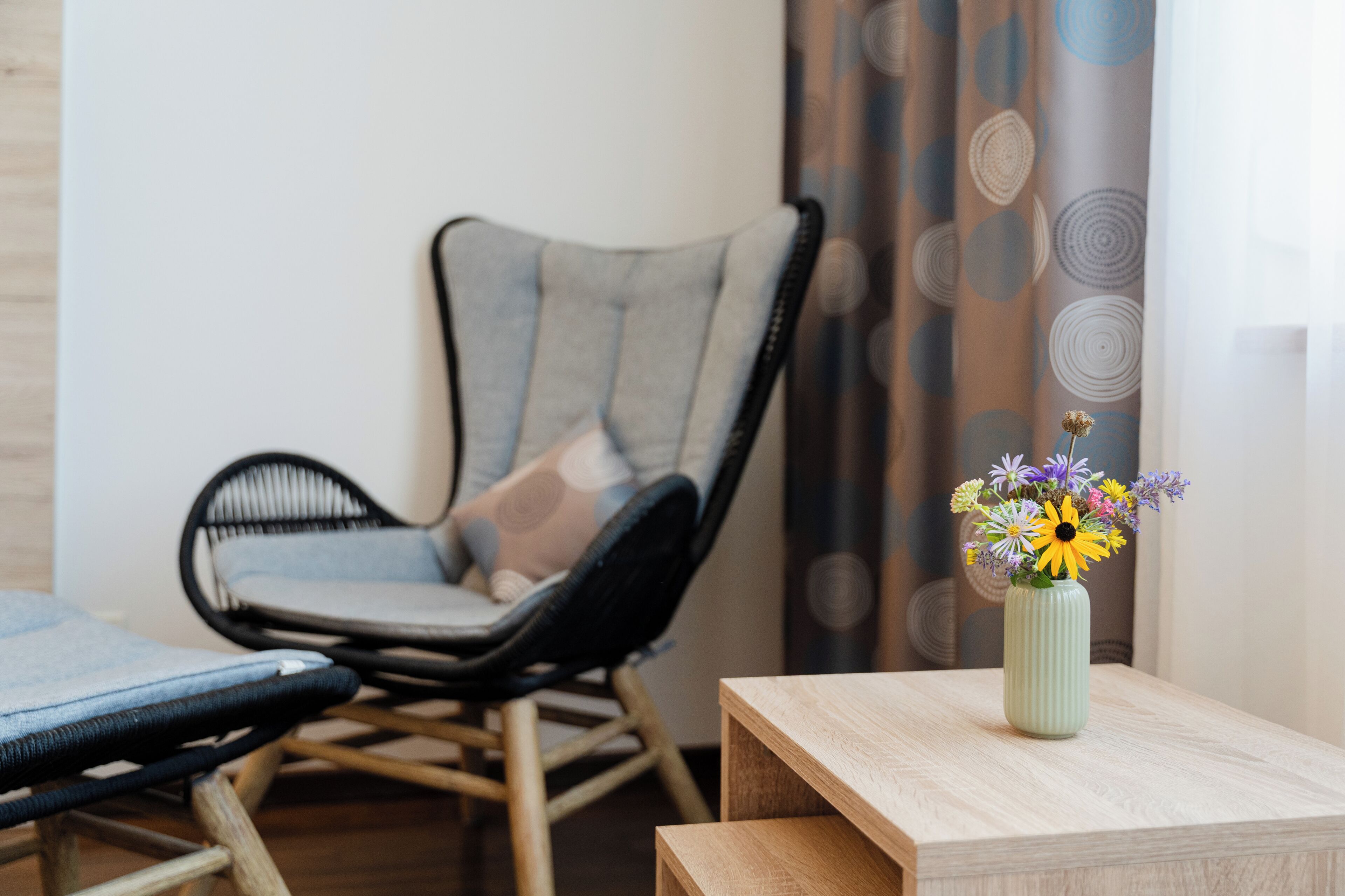 Une pièce avec une chaise en bois, une petite table sur laquelle est posé un vase de fleurs et un rideau à motifs.