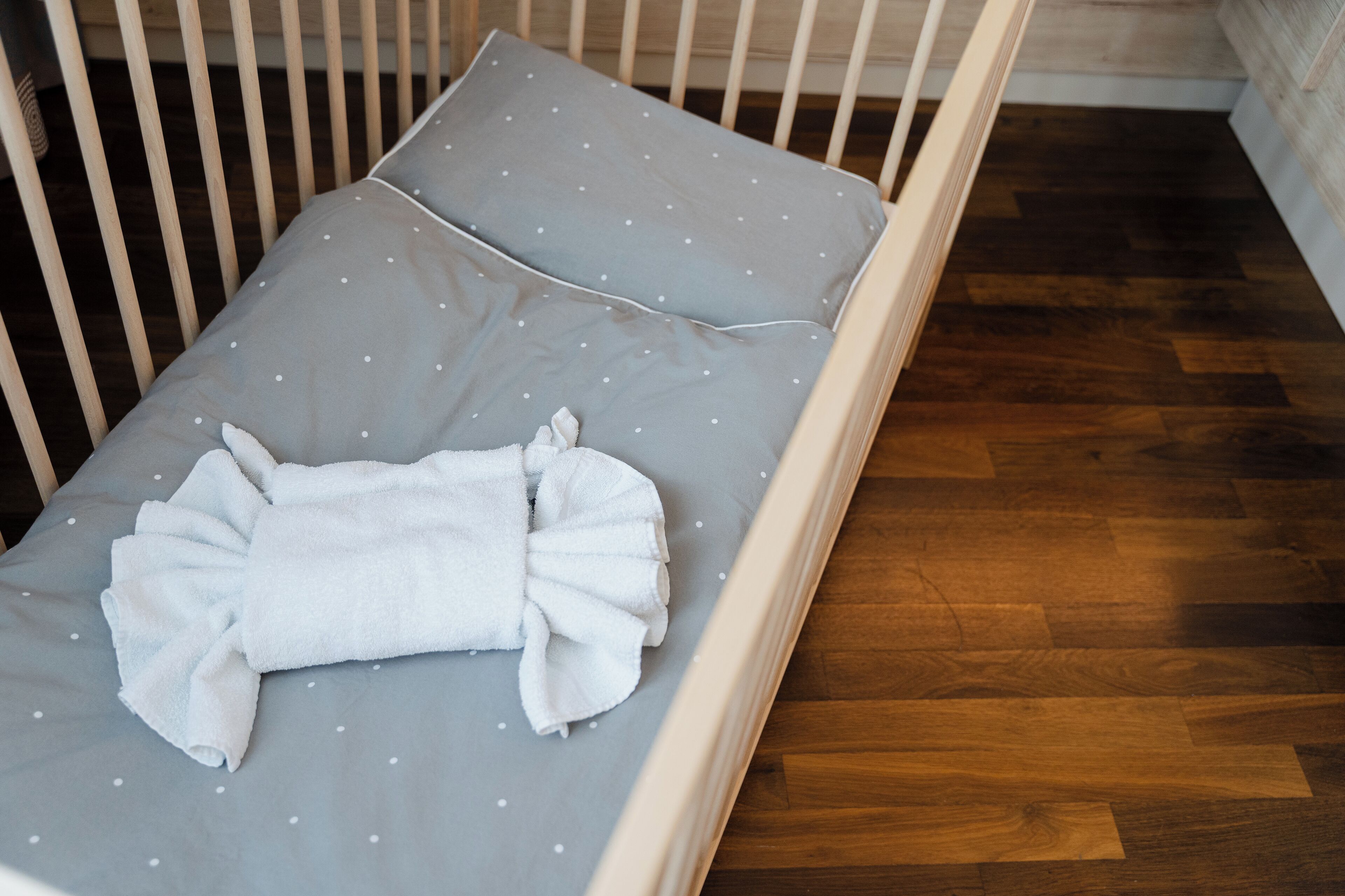 A crib with a grey mattress and white polka dots, a white blanket, and a wooden floor.