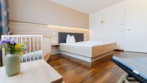 A modern bedroom with a wooden floor, a bed with a white mattress and gray headboard, a bedside table with a vase of flowers, and a crib in the corner. - Klostergasthof Roggenburg (Roggenburg)
