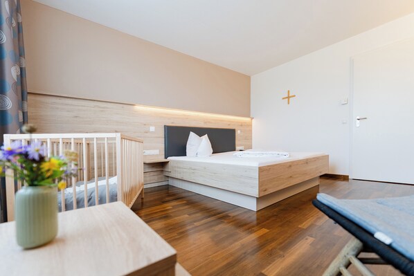 A modern bedroom with a wooden floor, a bed with a white mattress and gray headboard, a bedside table with a vase of flowers, and a crib in the corner.