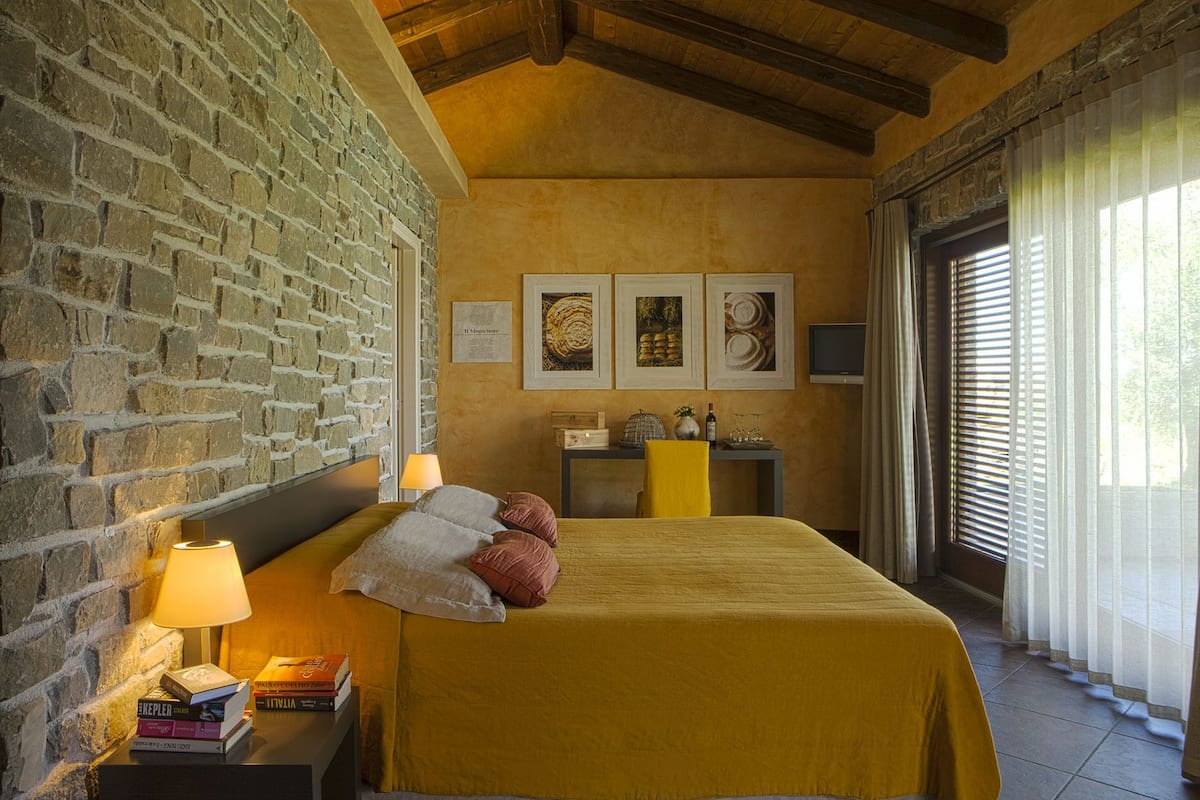A room with a bed, a nightstand with books, a lamp, a desk with a chair, and a window with curtains.