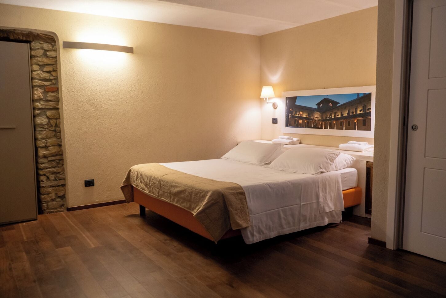 A hotel room with a bed, a stone wall, a nightstand with a lamp, and a painting on the wall.