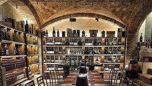 Una bodega con hileras de mesas y sillas de madera, estanterías llenas de botellas de vino y un techo abovedado.