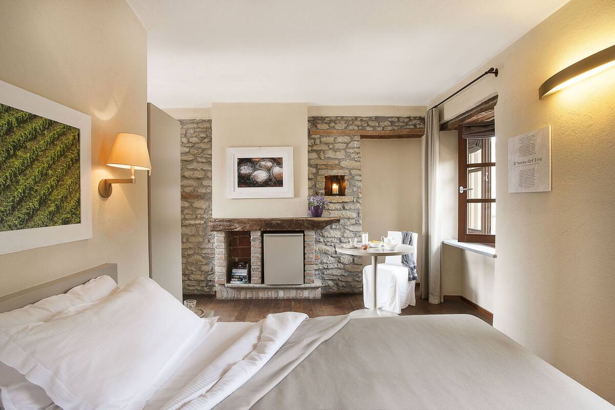 A hotel room with a bed, a stone fireplace, a small table with a vase, and a window with curtains.