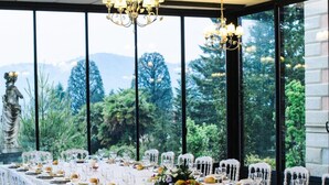 Una larga mesa de comedor con sillas blancas, una araña y vistas a la vegetación y montañas a través de amplios ventanales.