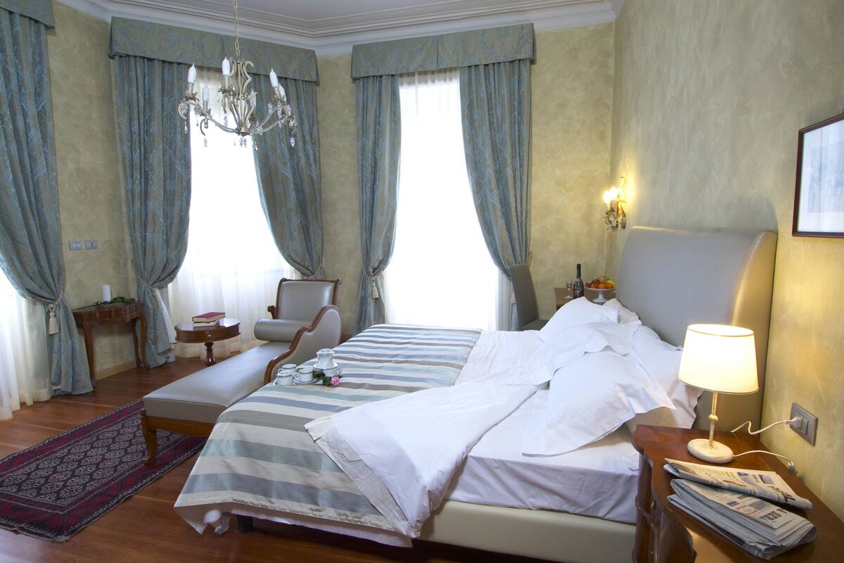 A hotel room with a large bed, a bedside table with a lamp, a chair, a small table with a vase and books, and a chandelier.