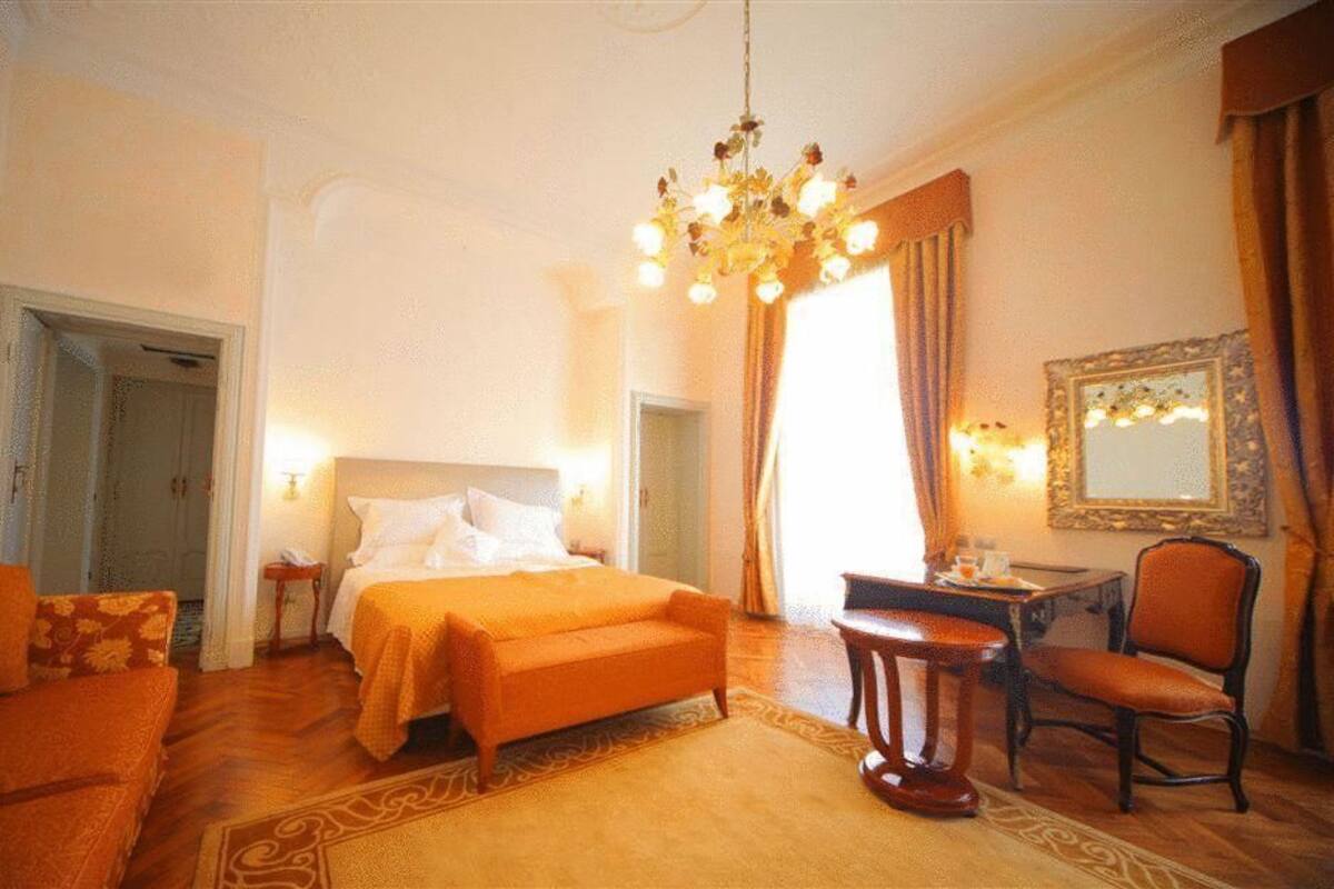 A hotel room with a bed, a desk, a chair, a chandelier, and a mirror.