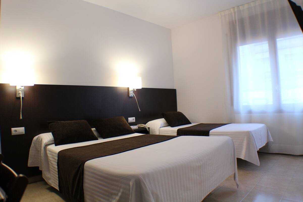 A hotel room with two beds, each with a headboard, white bedding, and a brown throw blanket. There is a window with sheer curtains and a wall-mounted light fixture.