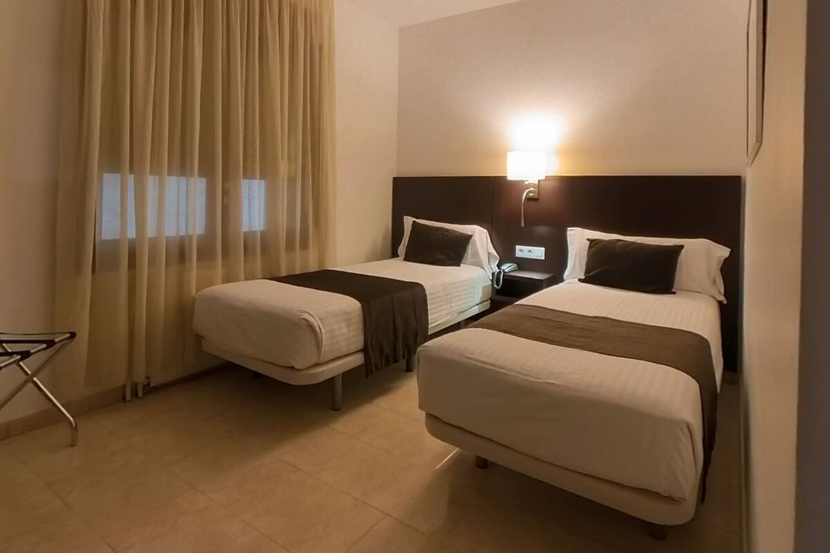 A hotel room with two beds, a desk, a chair, and a lamp.