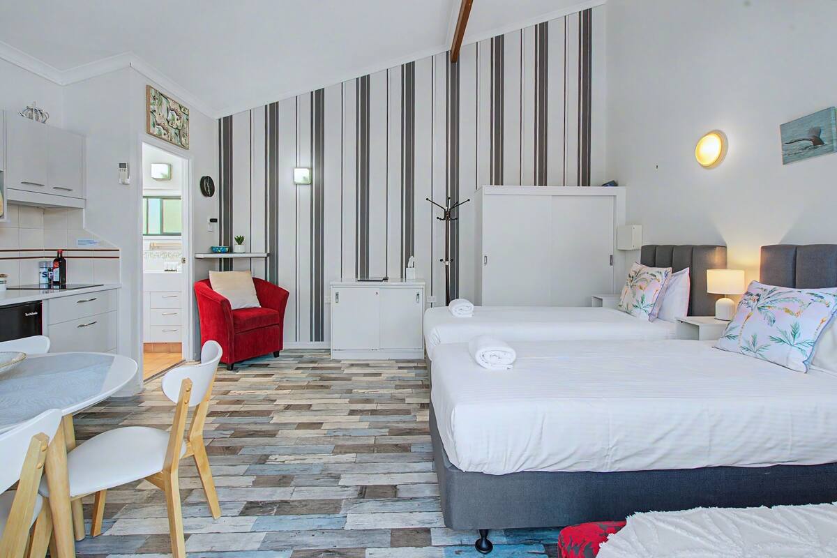 A hotel room with a kitchen area, a dining table with chairs, a red sofa, two beds with white linens, and striped wallpaper.