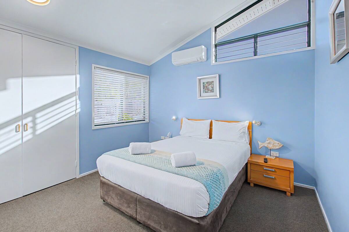 A bedroom with a bed, a nightstand, a window with blinds, an air conditioner, and a framed picture on the wall.