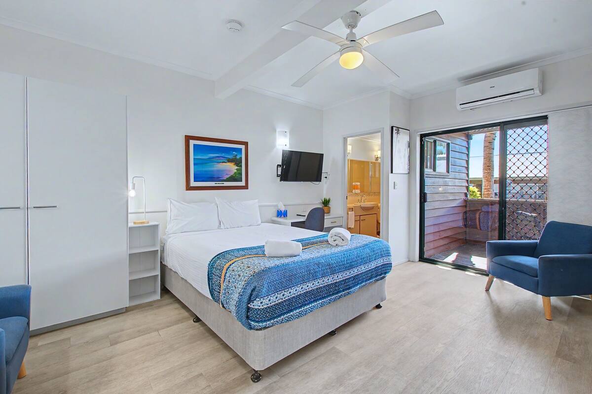 A hotel room with a bed, a ceiling fan, a TV, a sliding glass door to a balcony, and a blue chair.