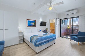 A hotel room with a bed, a ceiling fan, a TV, a sliding glass door to a balcony, and a blue chair. - Boat Harbour Stays (Urangan)