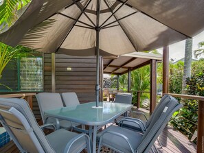 Relax in a shaded patio area surrounded by lush greenery and palm trees, with a glass-top table and comfortable seating.