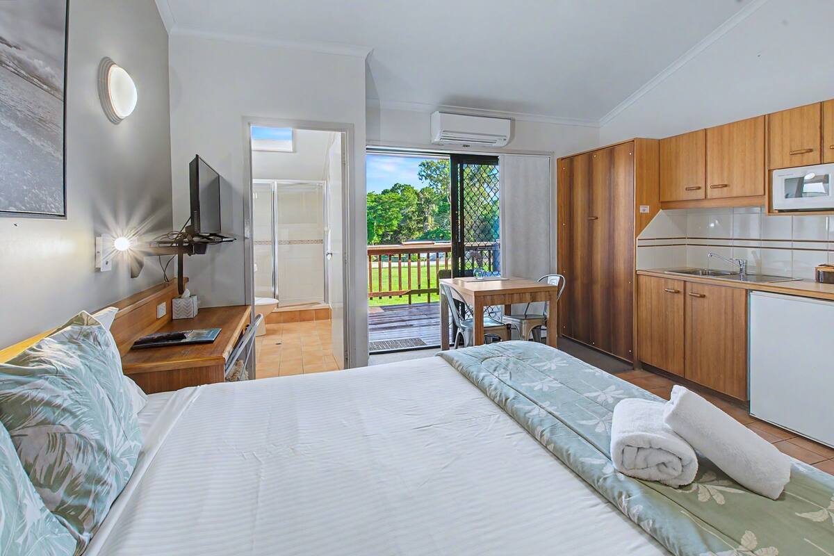 A bedroom with a bed, a desk, a chair, a TV, a kitchen area with cabinets and a microwave, and a sliding glass door to a balcony.
