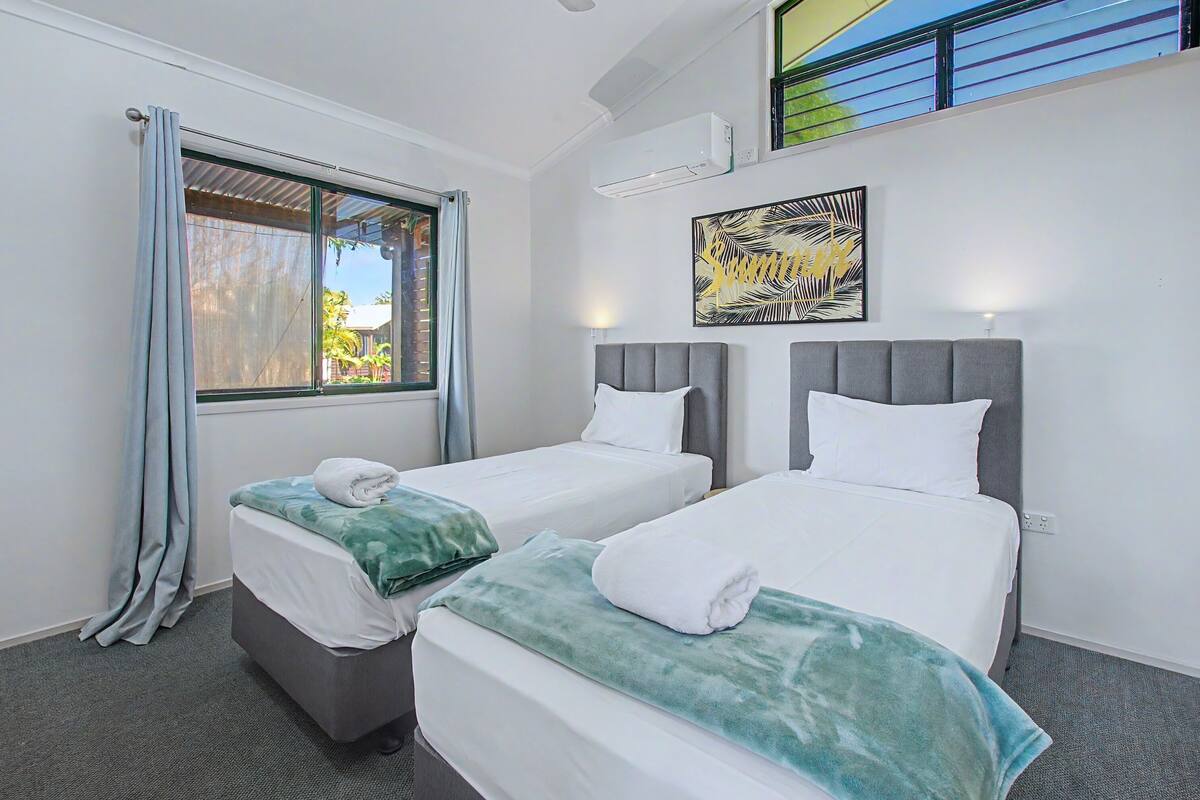 A hotel room with two beds, a window with curtains, an air conditioning unit, and a wall-mounted artwork.
