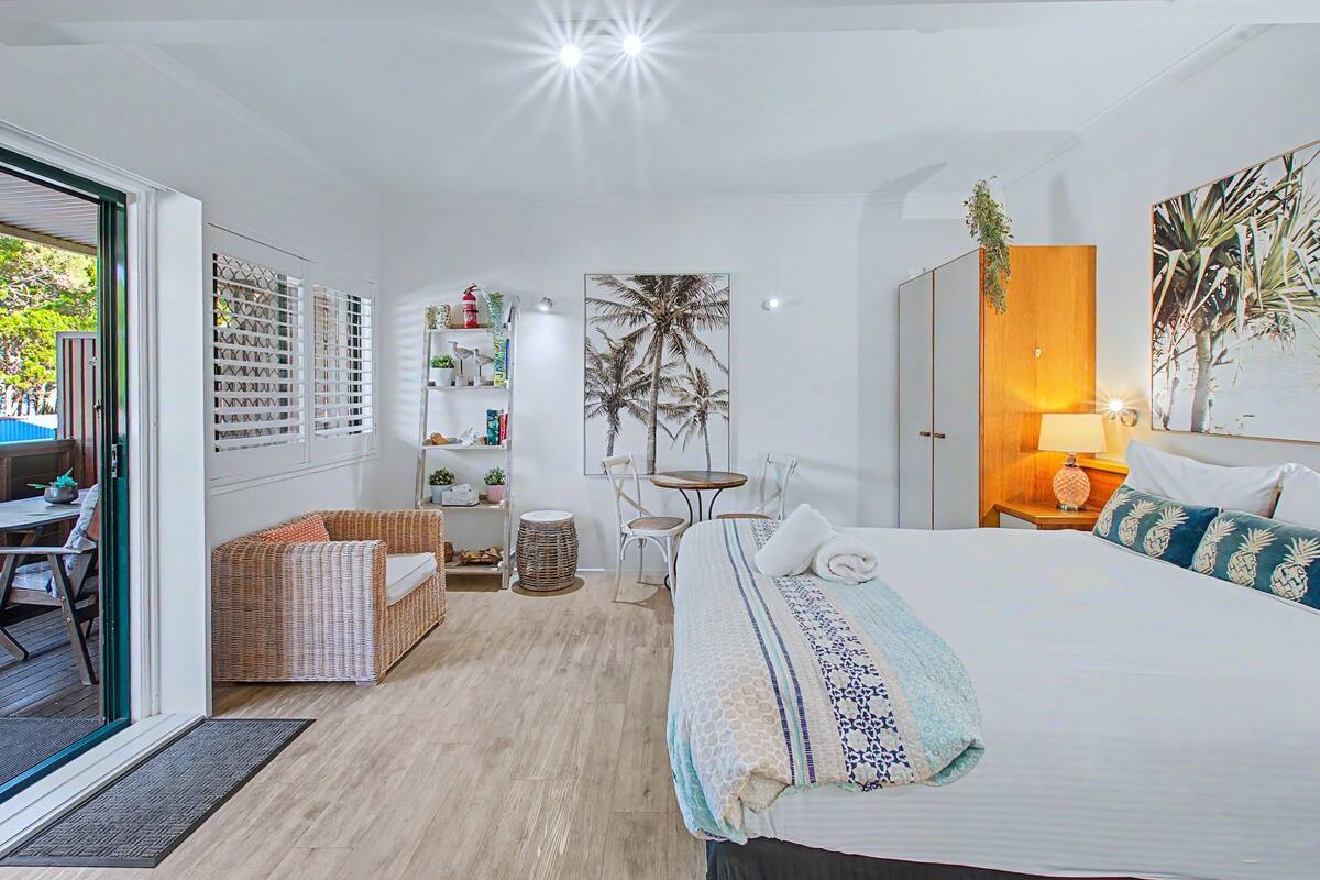 A hotel room with a bed, a wicker sofa, a small table with chairs, a bookshelf, and a large window.