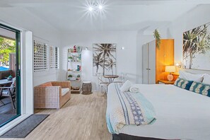 A hotel room with a bed, a wicker sofa, a small table with chairs, a bookshelf, and a large window. - Boat Harbour Apartments (Urangan)