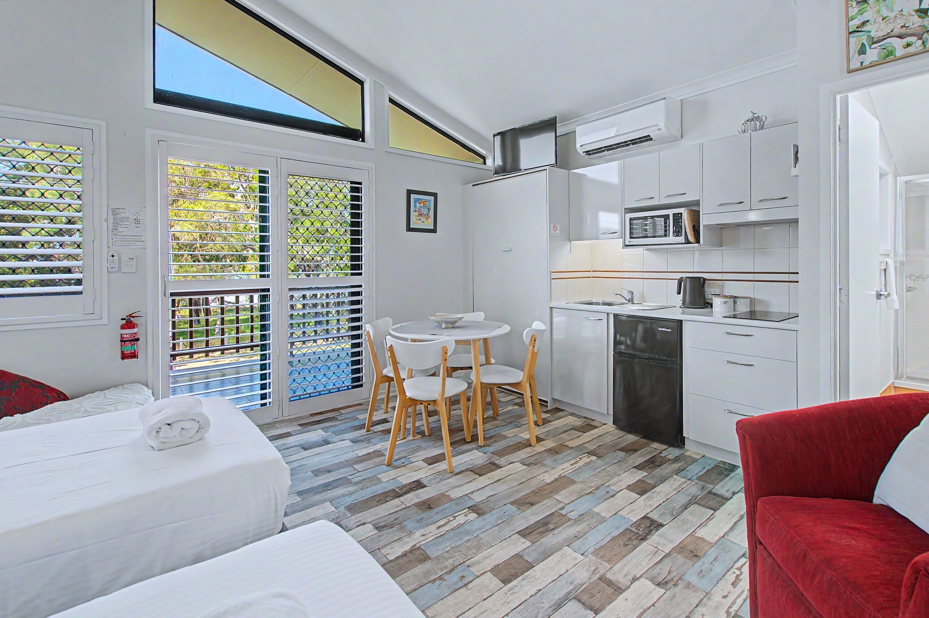 Una habitación de hotel compacta con cama, una pequeña mesa de comedor con sillas, kitchenette y un sofá rojo.