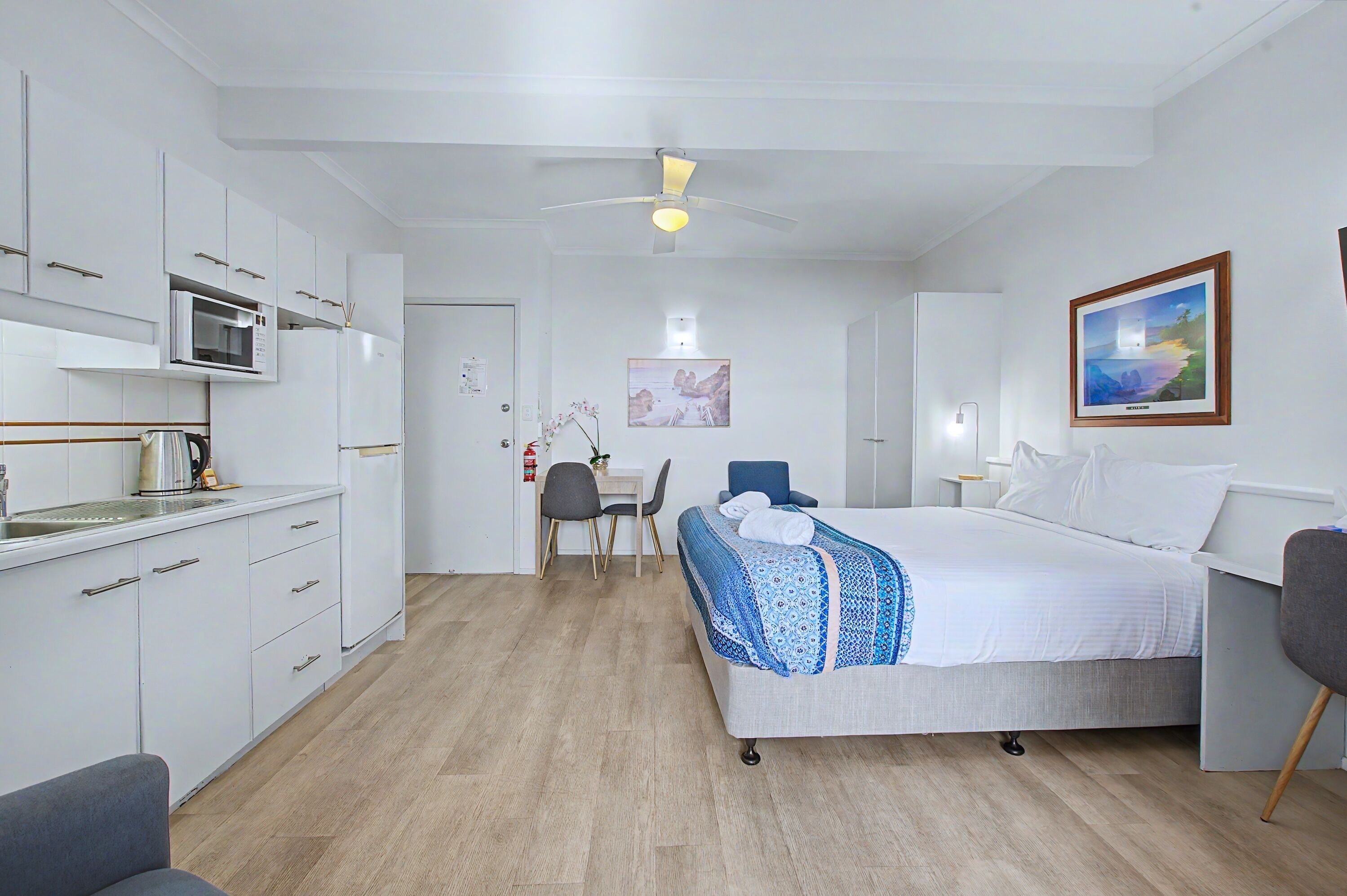 Una habitación de hotel moderna con kitchenette, una cama con colcha azul y blanca estampada, un escritorio con silla y un cuadro en la pared.