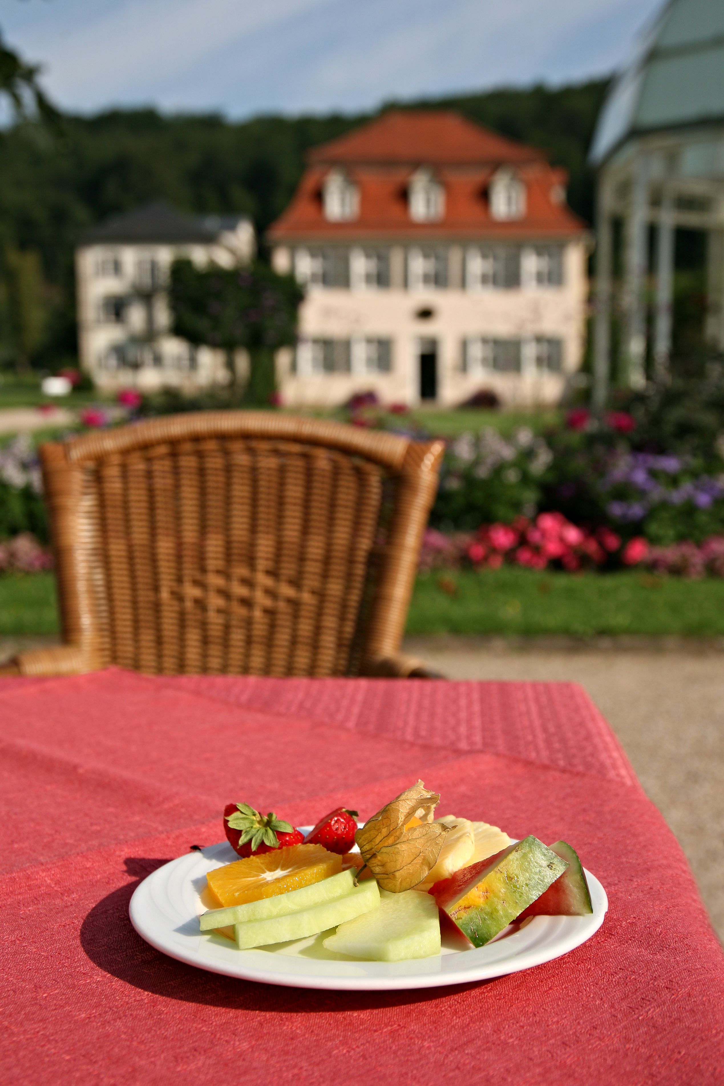 A plate of fruit on a red tablecloth with a wicker chair and a building in the background.