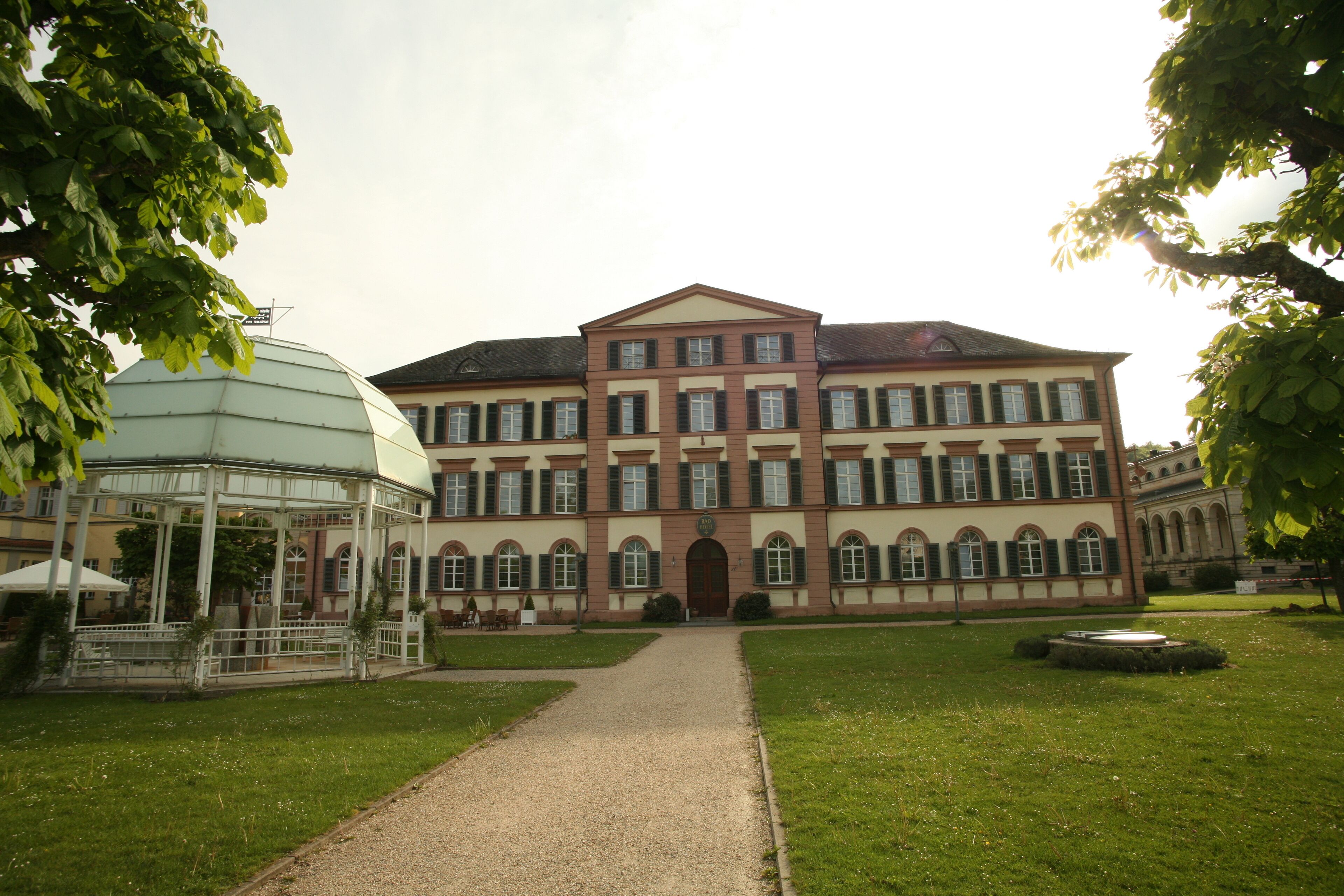 Ein großes, historisches Gebäude mit einem zentralen Eingang, umgeben von gepflegten Rasenflächen und einem überdachten Pavillon.