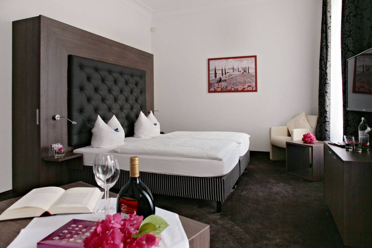 Une chambre d’hôtel avec un grand lit, une table de chevet sur laquelle se trouve une bouteille de vin, un livre ouvert et un bouquet de fleurs.