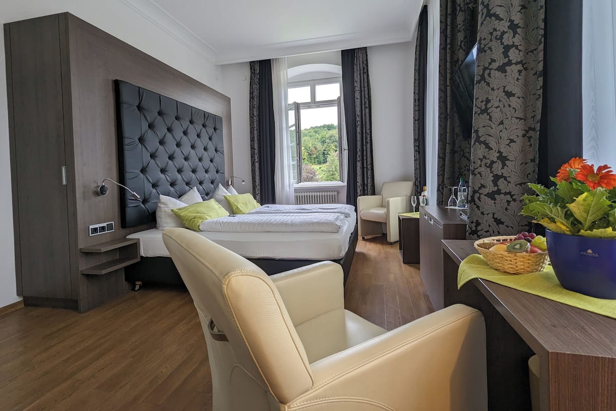 Une chambre d’hôtel dotée d’un grand lit, d’un coin salon, d’un bureau agrémenté d’un vase de fleurs et offrant une vue sur la verdure environnante.
